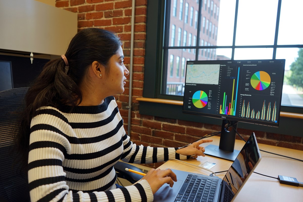 A young woman in a black and white striped shirt looks at a monitor while working on a laptop while sitting at a desk near a window.