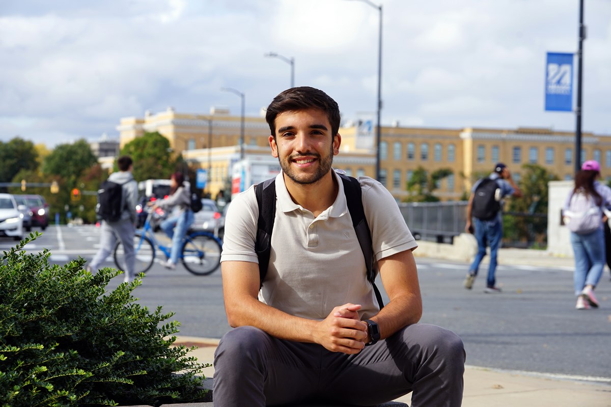 A young man in a white polo shirt wearing a backpack poses for a photo in front of an intersection where people are walking and biking.