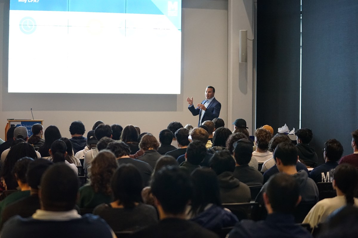 A man in a blazer speaks to a room full of people while standing in front of a projection screen.