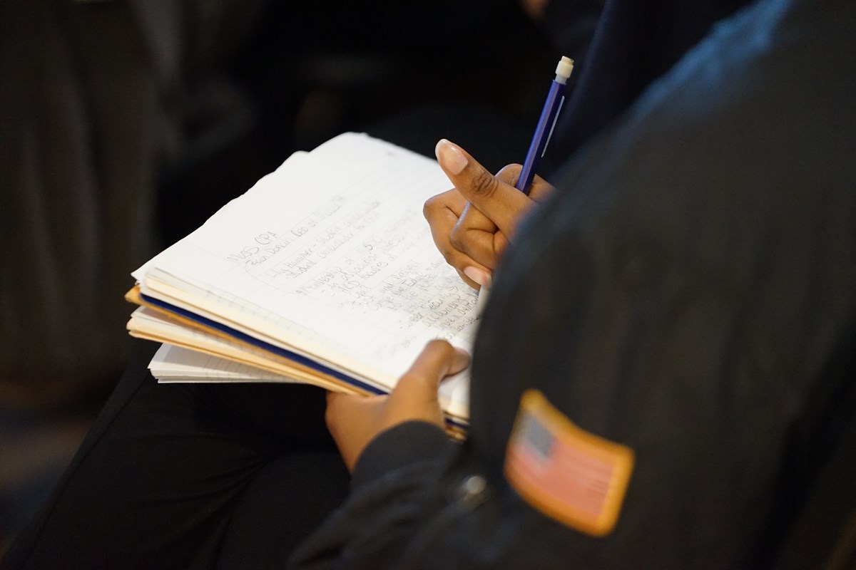 A person takes notes on a spiral notebook while sitting.