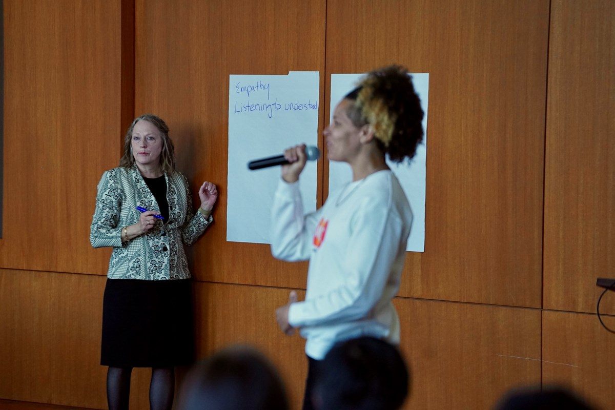 A woman speaks into a microphone while in the background a woman waits to write on a large piece of paper that is taped to the wall.