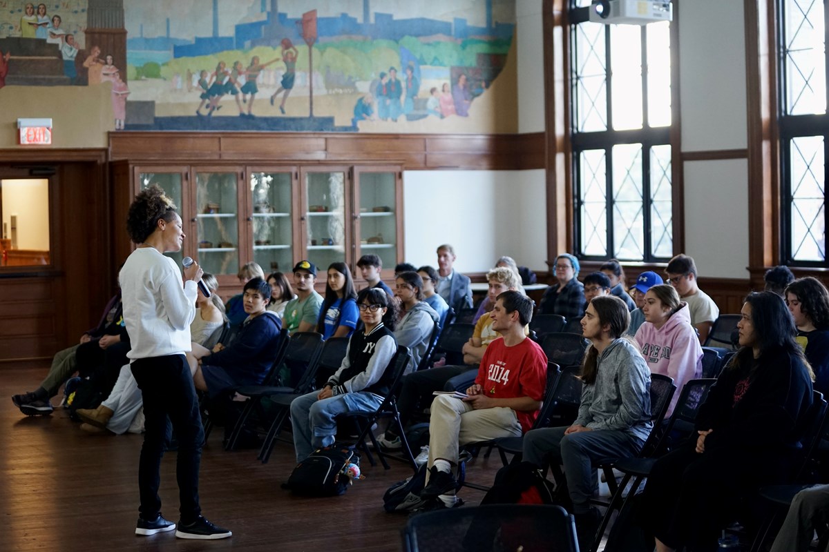 A woman in a white sweatshirt and black pants holds a microphone while speaking to an audience in a room.