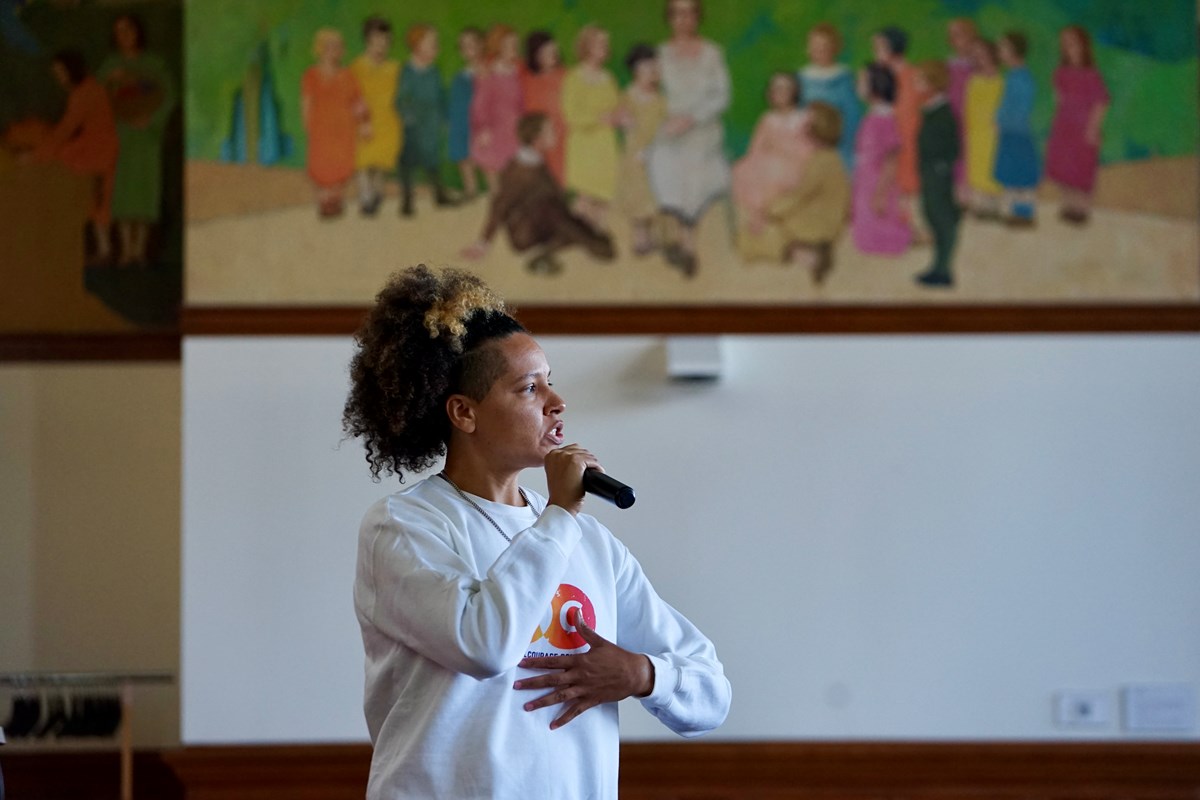 A woman speaks into a microphone while standing in a room where there is a colorful mural of school kids painted on the wall behind her.