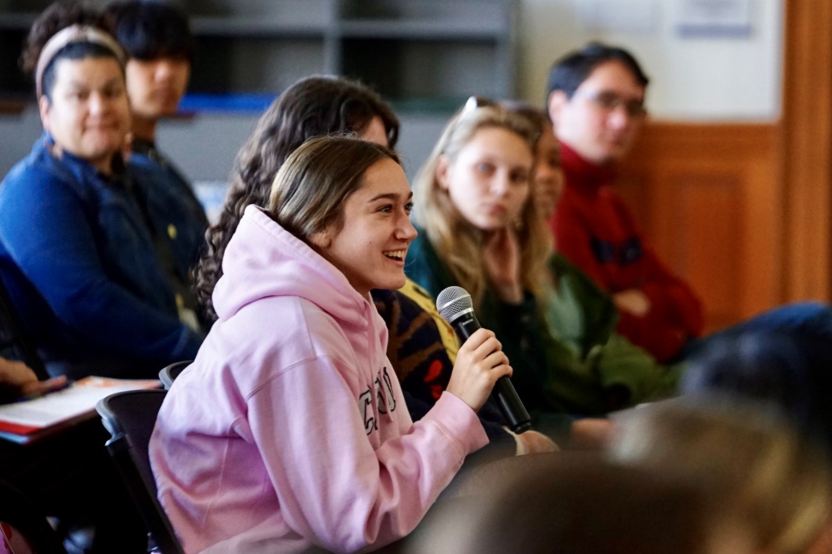 A young person in a pink hoodie smiles while speaking into a microphone while sitting in an audience.