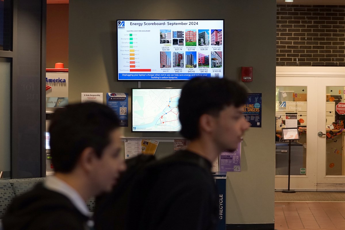 Two young people walk past a monitor on a wall in the lobby of a college residence hall.