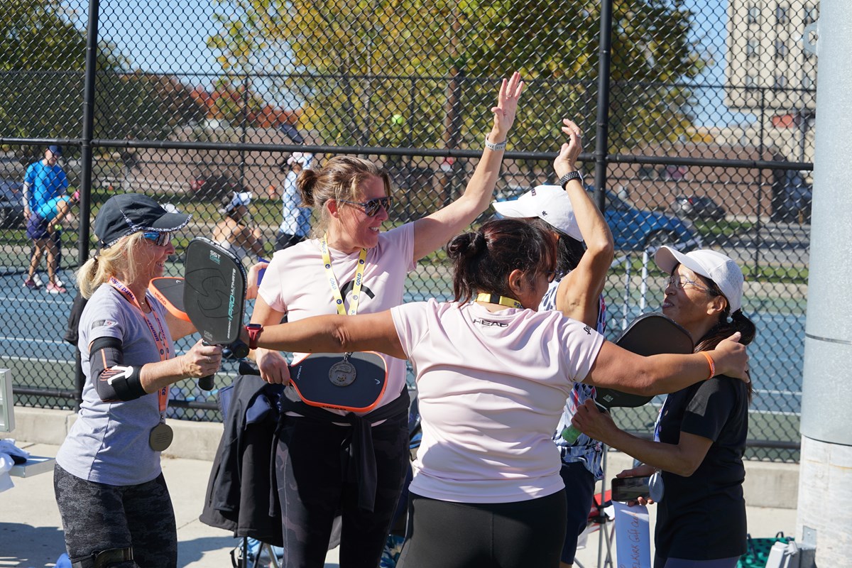 Five women celebrate near a pickleball court outdoors.