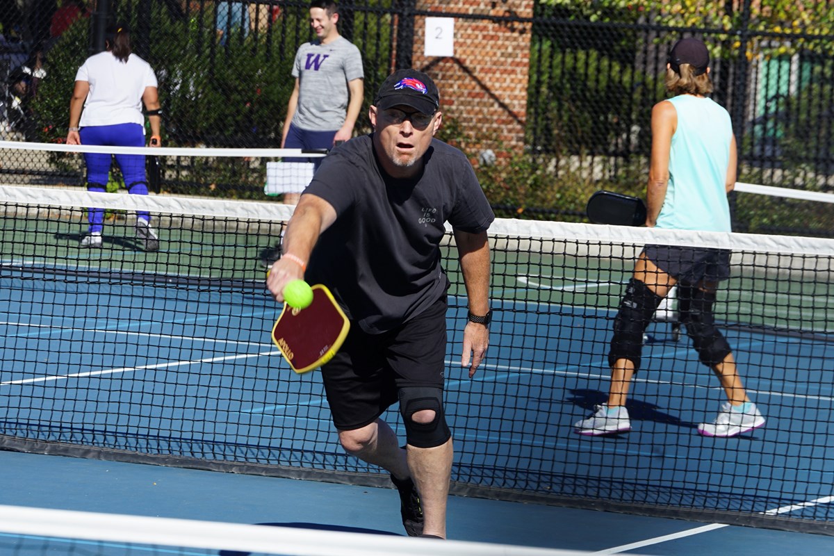 A man in dark shorts, a T-shirt and hat lunges for a shot during a game of pickleball.