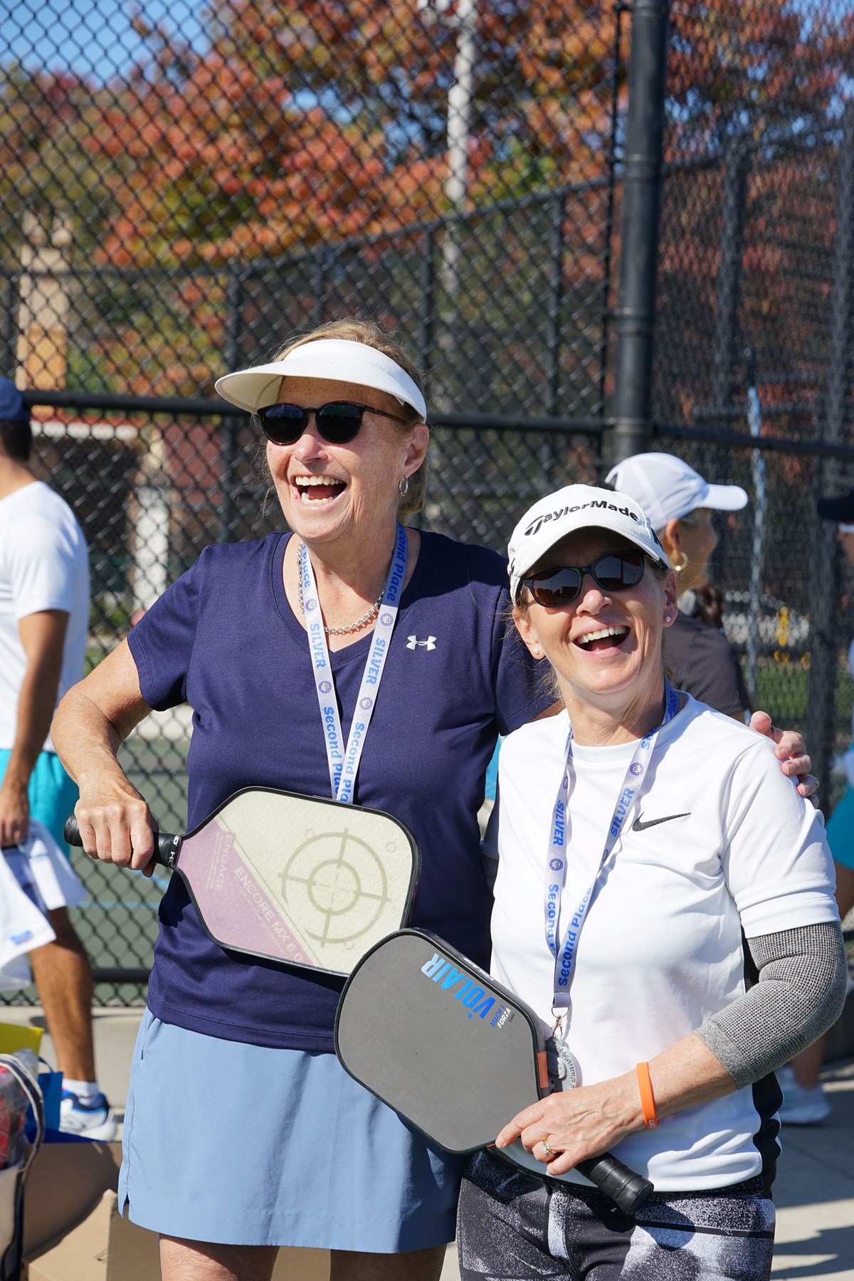 Two women in visors and sun glasses smile while holding pickleball paddles outside.