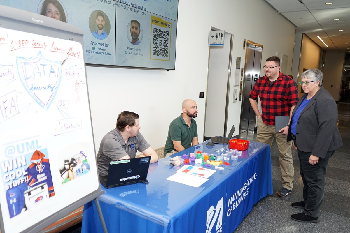 Two people are standing and talking to two other people who are seated behind a table with a blue tablecloth in the lobby of a building.