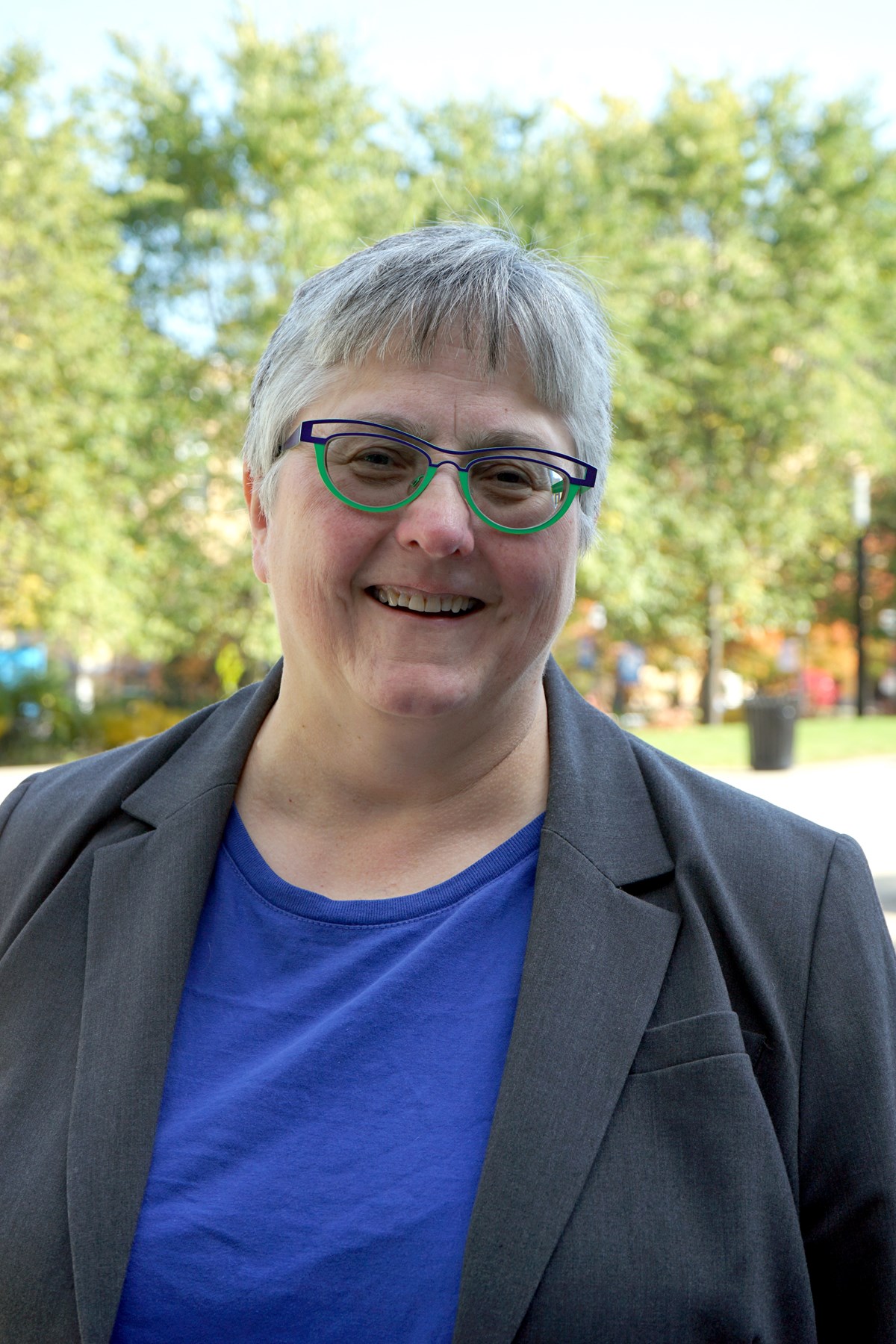 A person wearing glasses and a blazer poses for a photo outdoors.