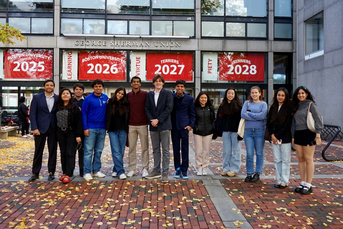 Thirteen young people stand in a line, posing for a photo outside of a college's student union.