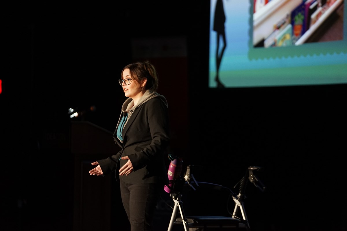 A person with dark hair and glasses makes a presentation on a stage. There is a walker next to her and a projection screen behind her.