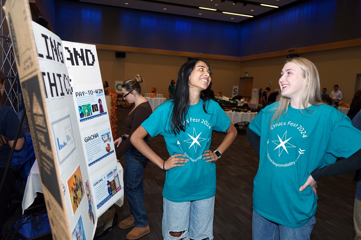 Two smiling young women in matching teal T-shirts stand with their hands on their hips in front of a poster board in a room.