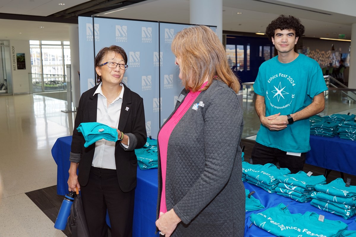 A woman with glasses holds a T-shirt while talking to a woman as a young man looks on.