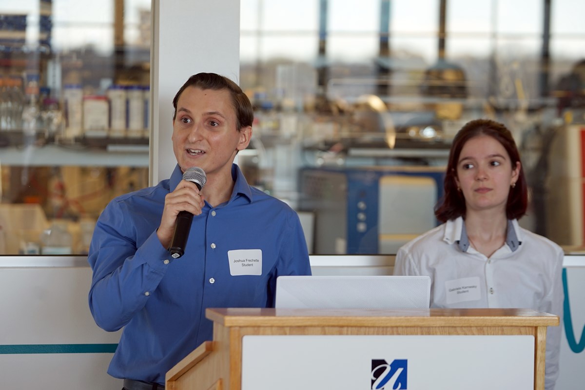 A student in a blue shirt holds a microphone and talks at a podium while another student looks on.