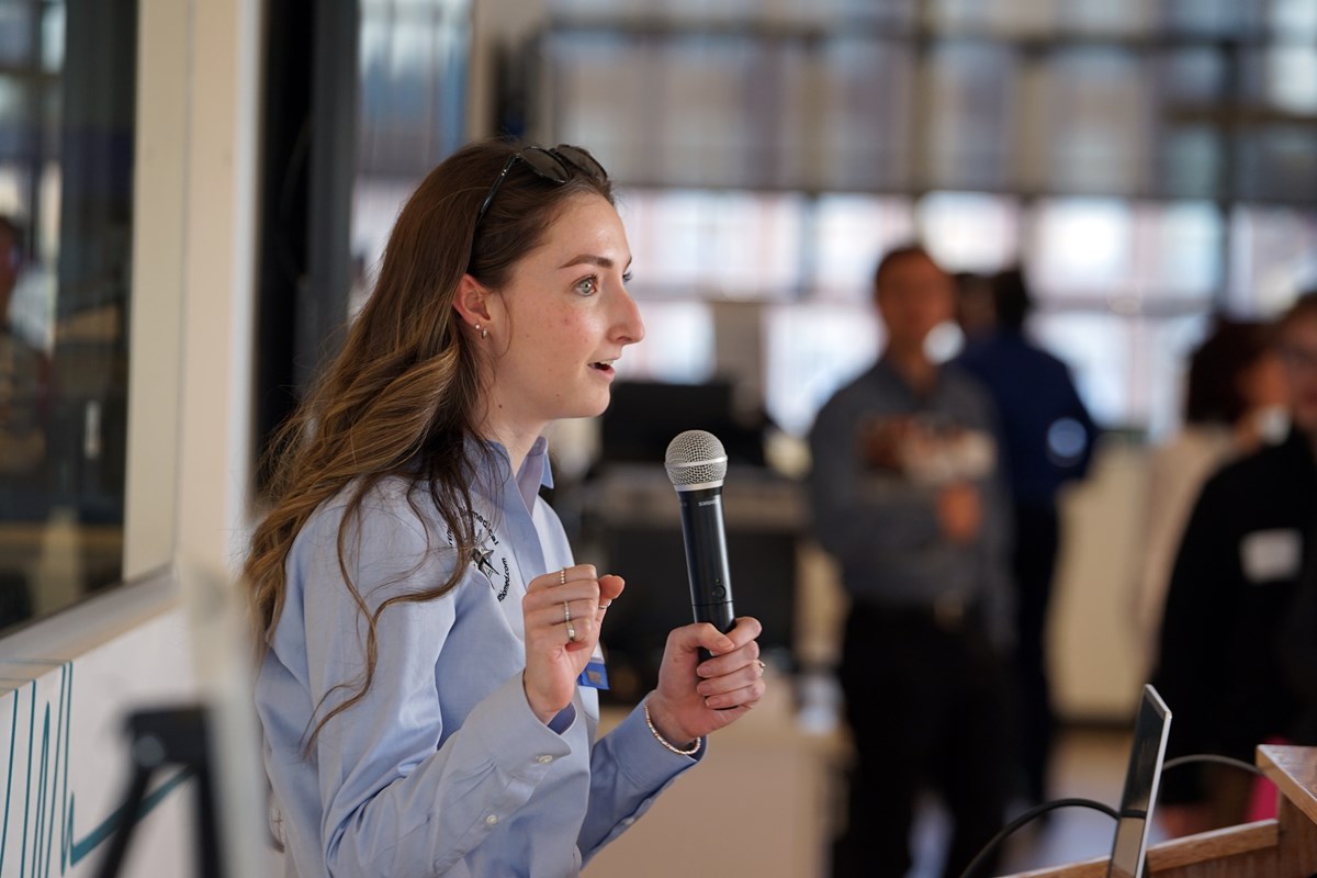 A young woman with long hair in a blue shirt holds a microphone while talking to a crowd in a room.