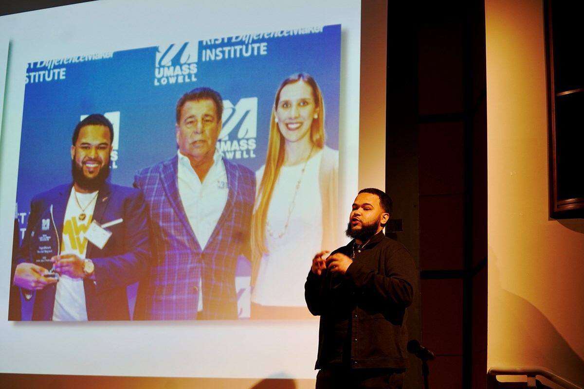 A young man in a dark suit presents on a stage in front of a projection screen.