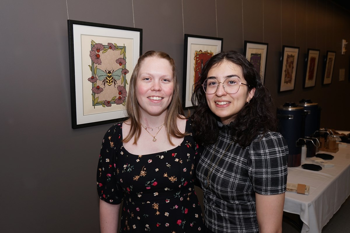 Two young women pose for a photo standing in front of a wall with framed illustrations of nature.