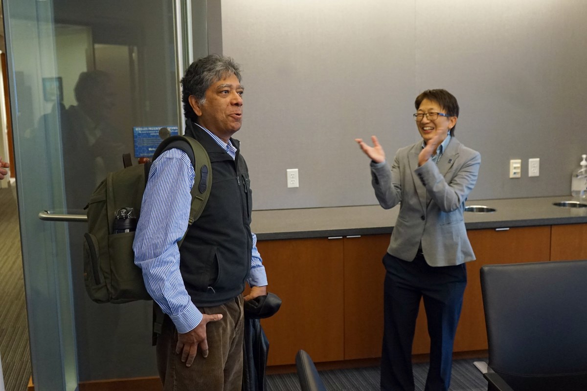 A man in a fleece vests looks surprised while walking in a room. A smiling woman claps as he enters.