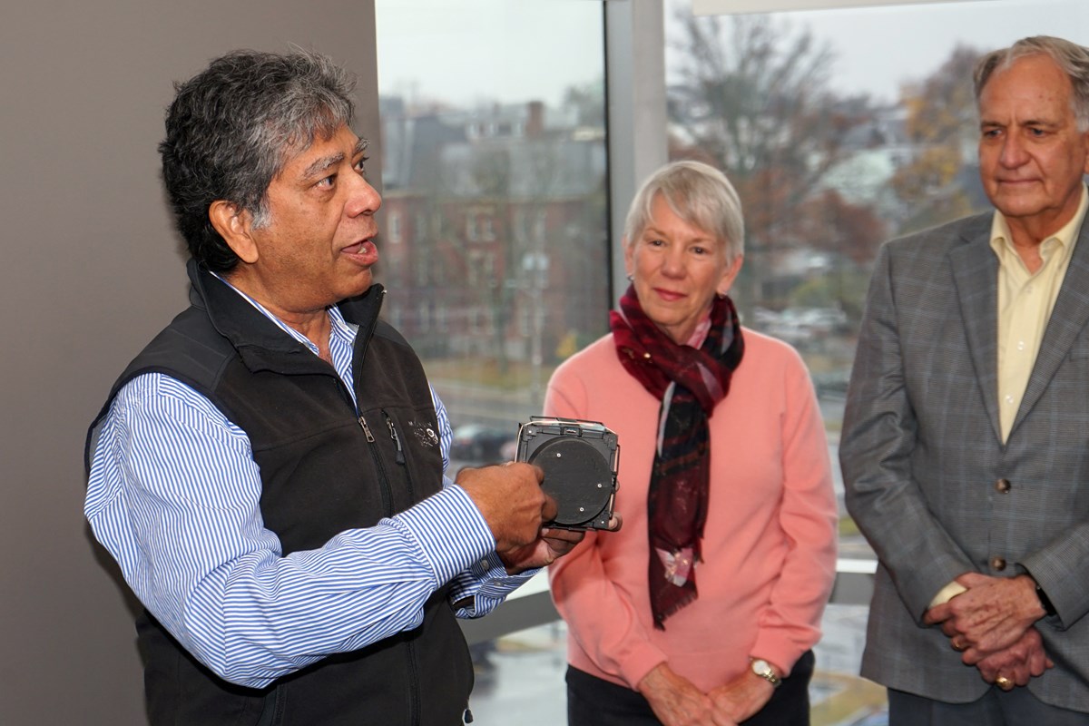 A man in a fleece vests points to a small device that he is holding while two people look on.