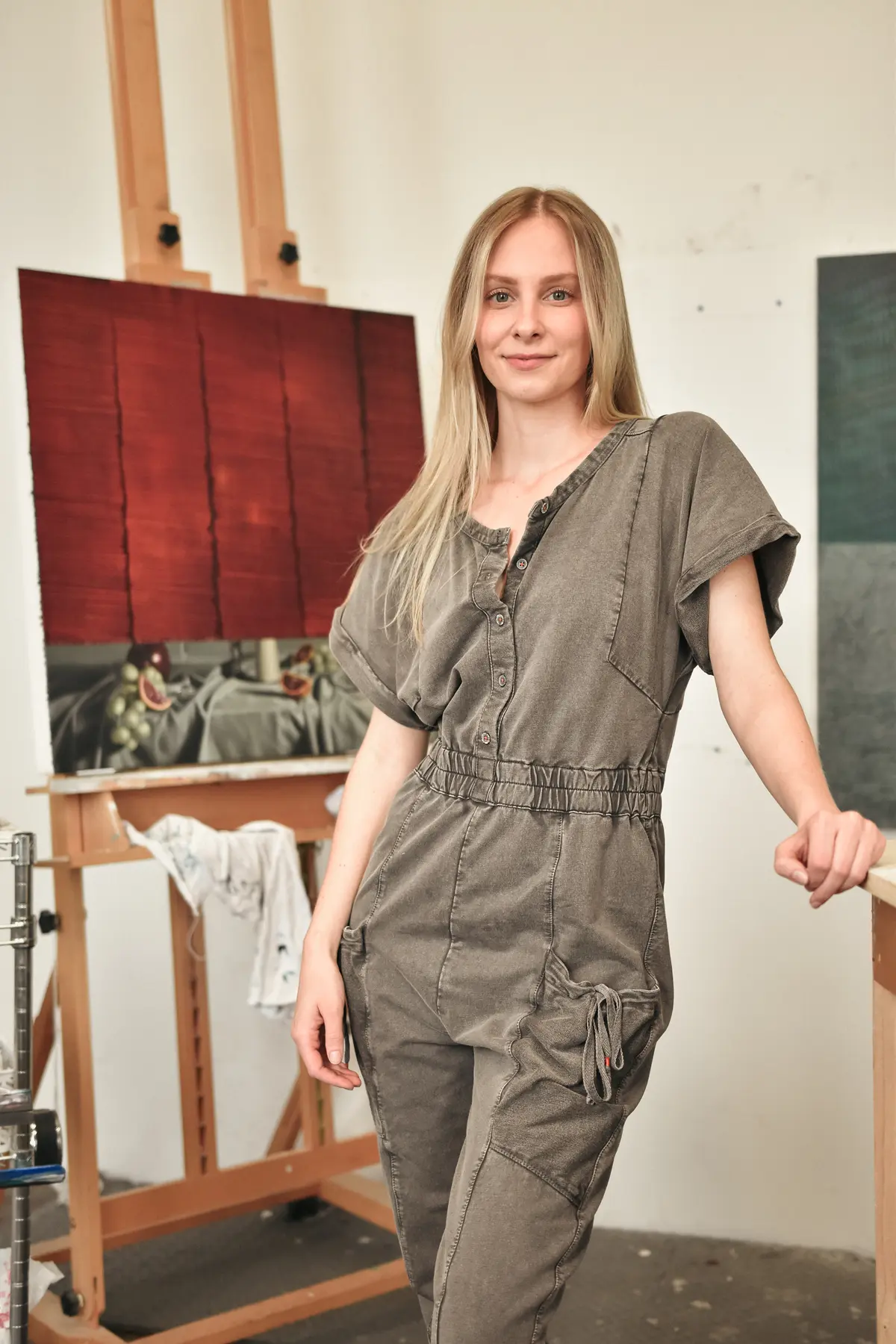 A young woman wearing a grey jumper poses for a photo next to an oil painting on an easle.