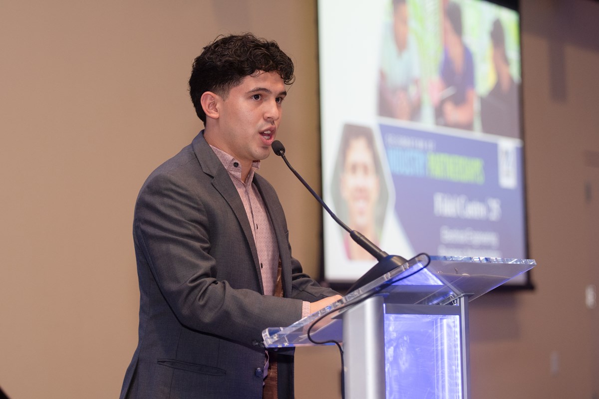 A young man speaks into a microphone while standing on a stage in front of a projection screen.