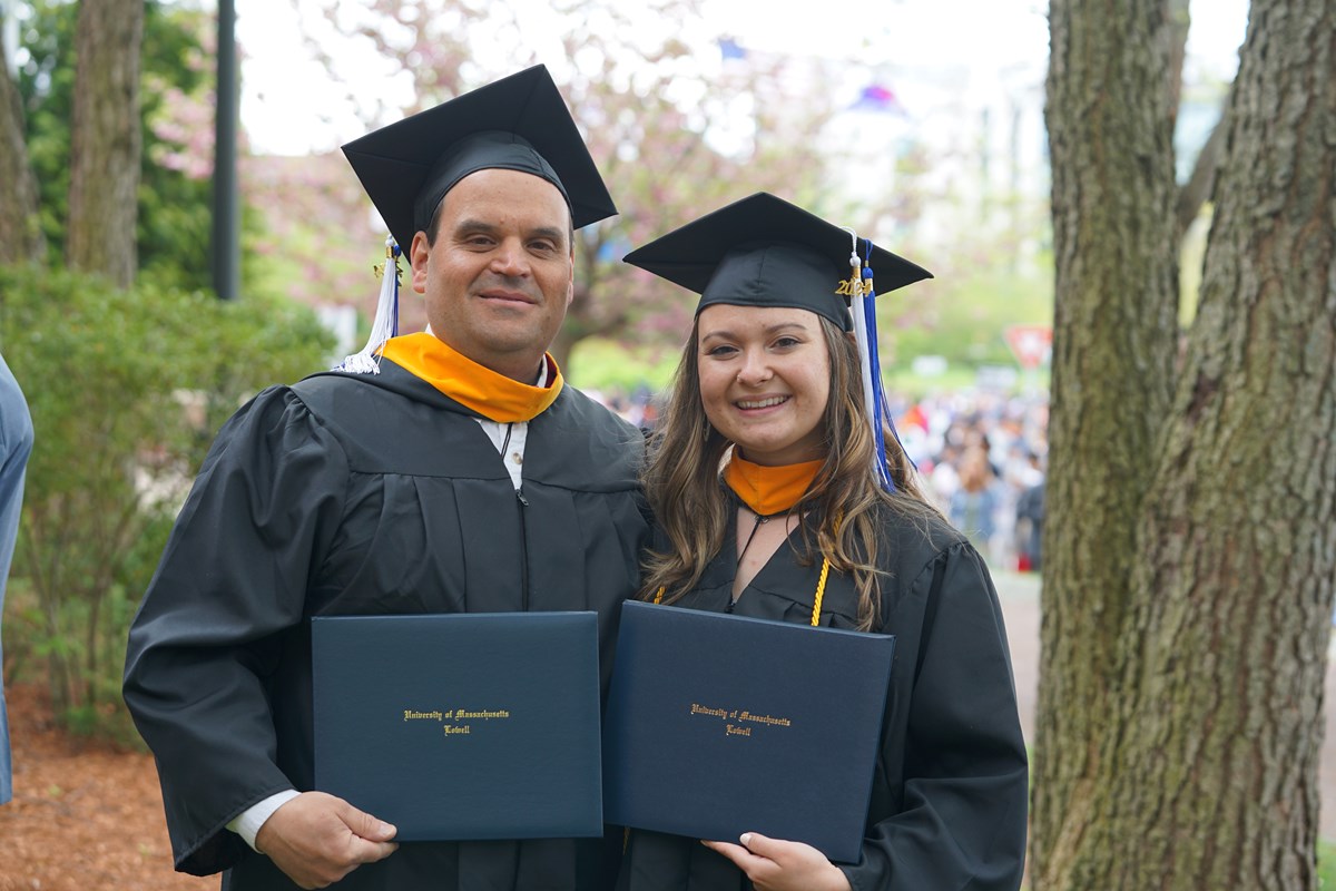 Two people pose for a photo outside in their graduation caps and gowns, holding diplomas.