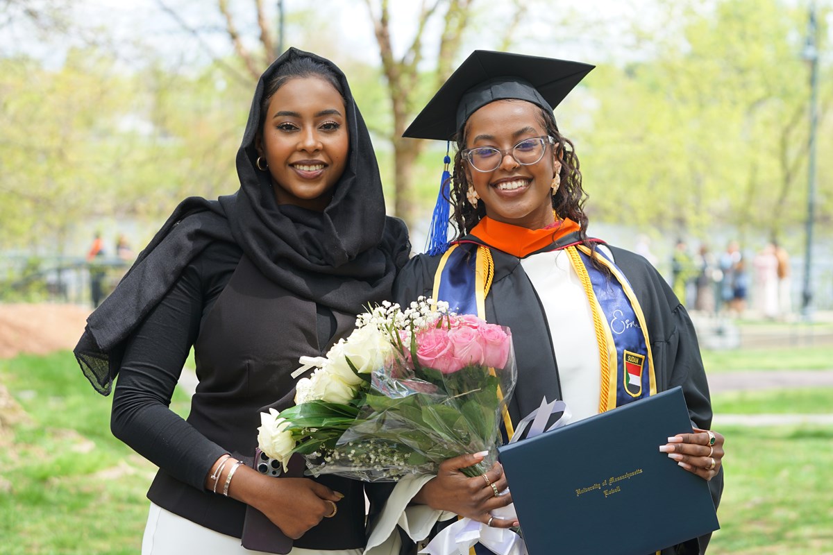 A person in a cap and gown holds flowers and a diploma while standing next to another person and posing for a photo outside.