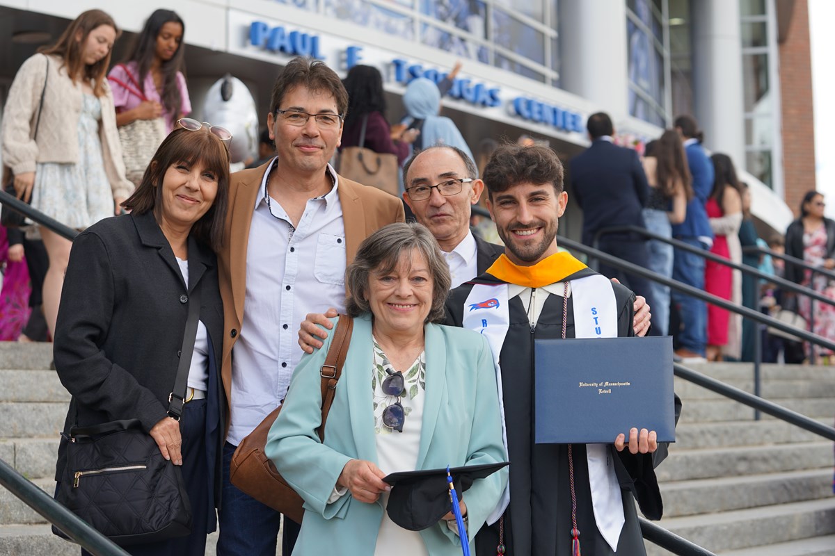 A person in a graduation gown holds his diploma while posing for a photo with four people on the steps outside an arena.