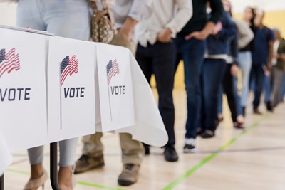 People stand in line to vote. A table has signs with flags and the word "vote."