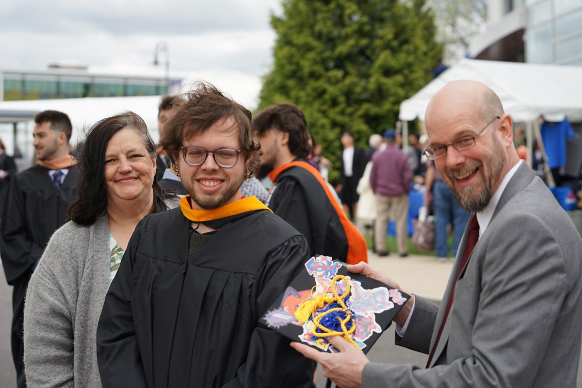 A person in a graduation gown poses for a photo outside while standing next to two people. One his holding his mortar board.