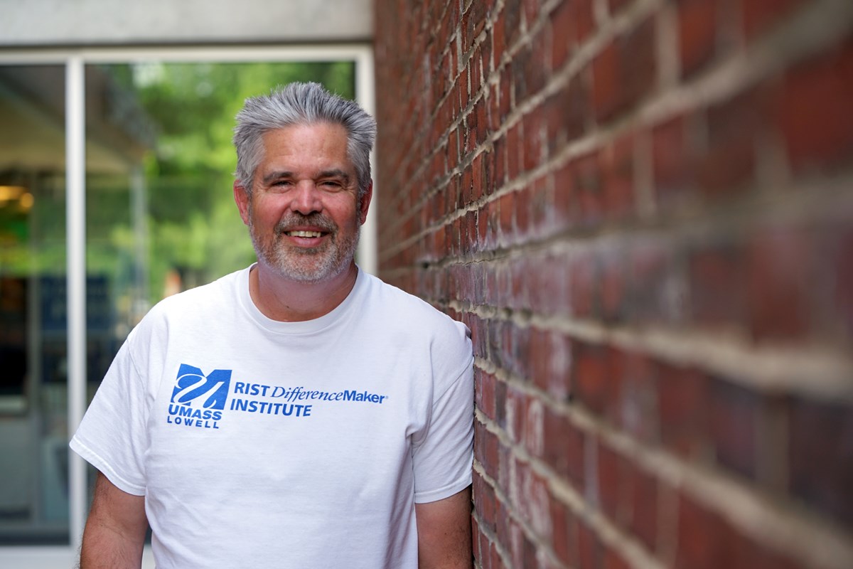A person with gray hair and a beard wearing a white T-shirt leans against a brick wall and poses for a photo.