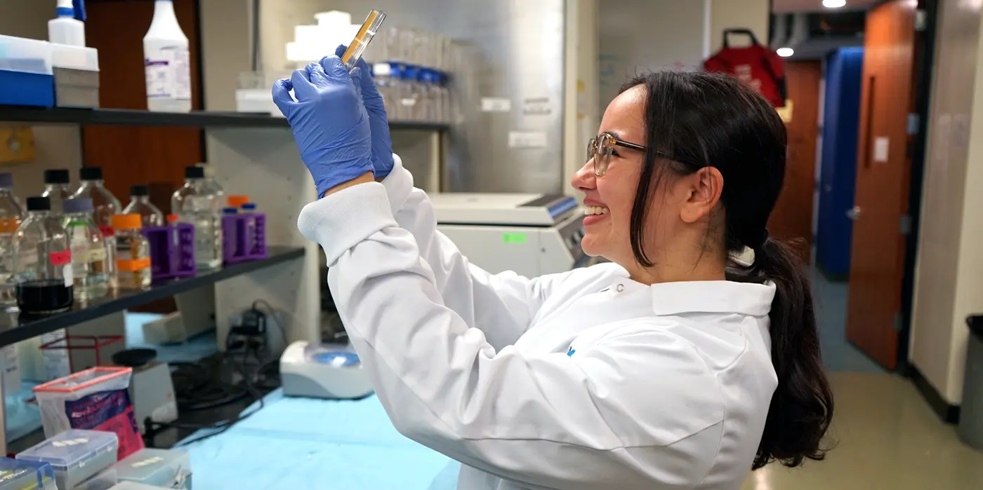 Student in a white labcoat smiles while examining the contents of a glass test tube in a UMass Lowell laboratory 