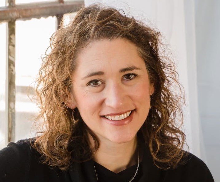 Caucasian woman with curly brown hairs in black shirt smiles at camera