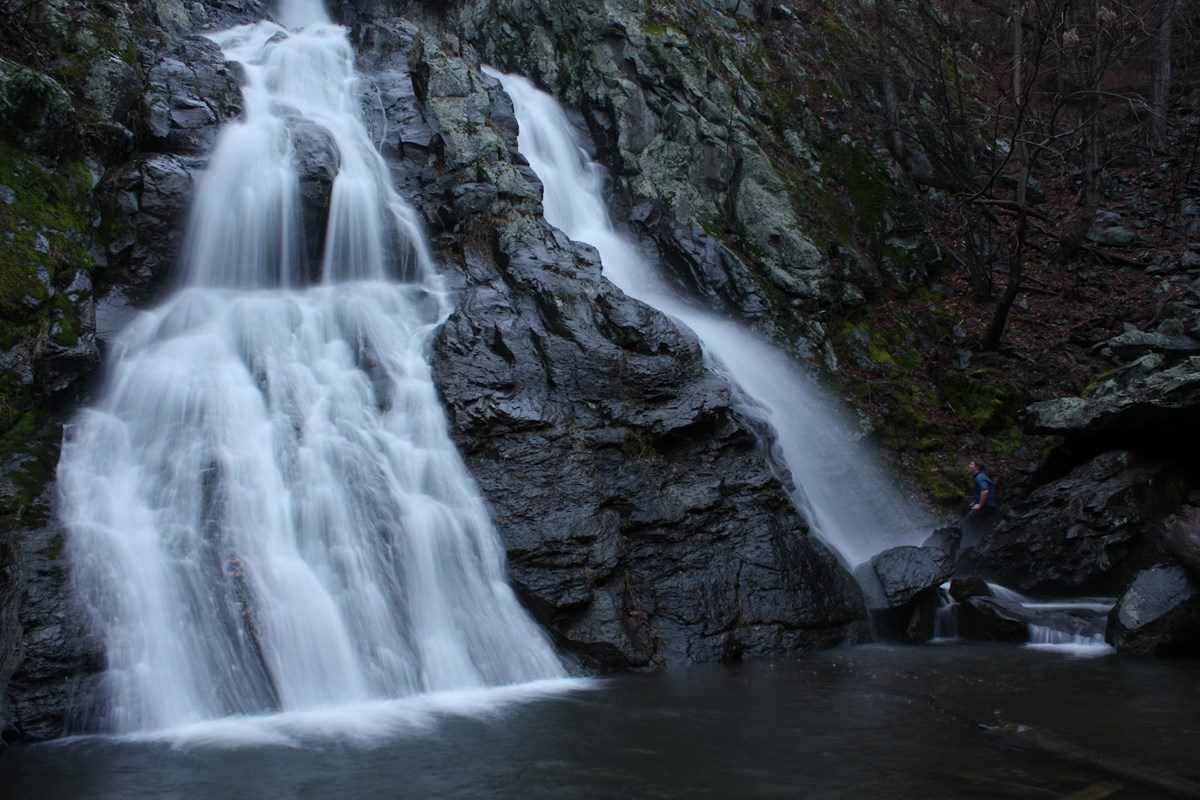 2 branches of a waterfall flows down a rock wall into a dark pool below.