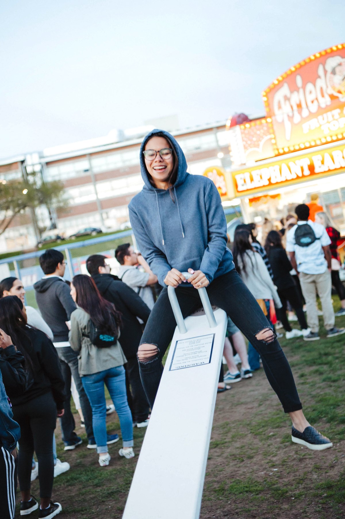 Student in hoodie on a seesaw with a carnival behind them.