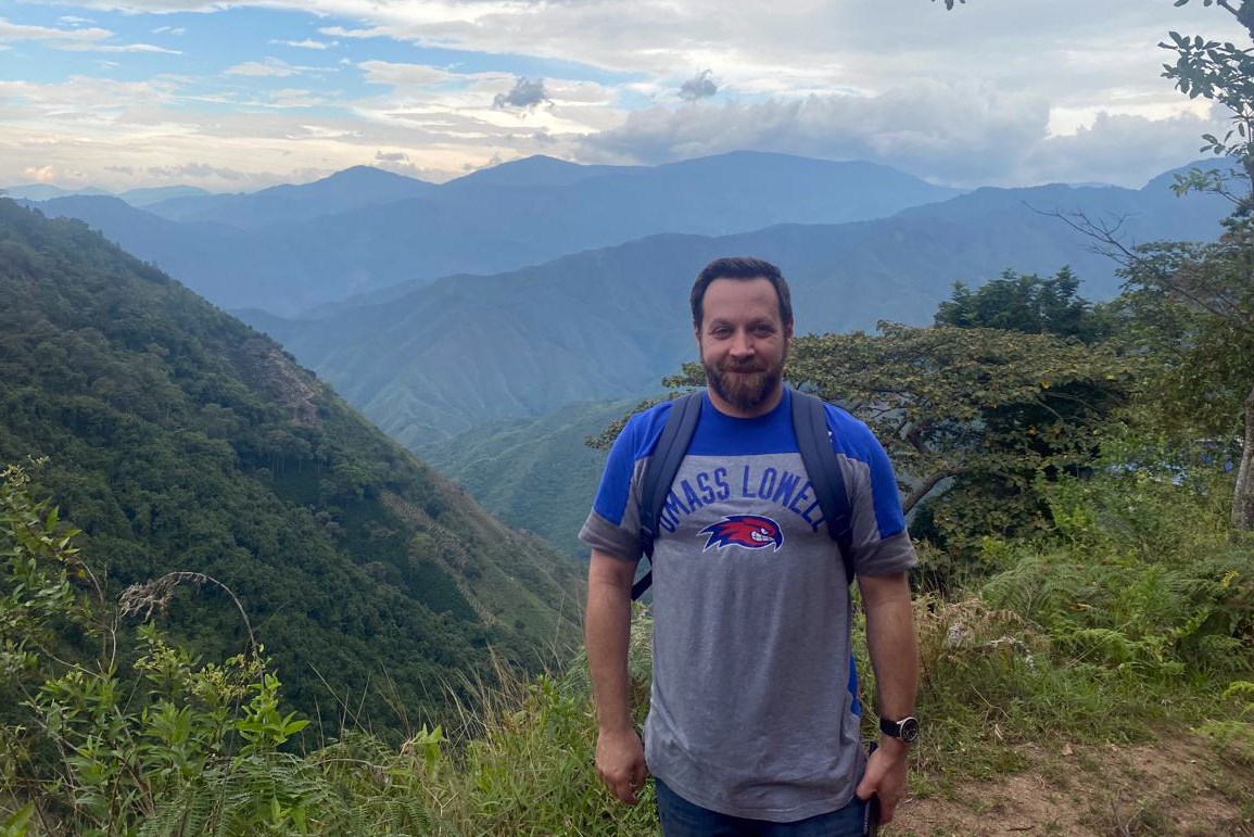A person with a beard wearing a backpack poses for a photo in front of mountainous valley.