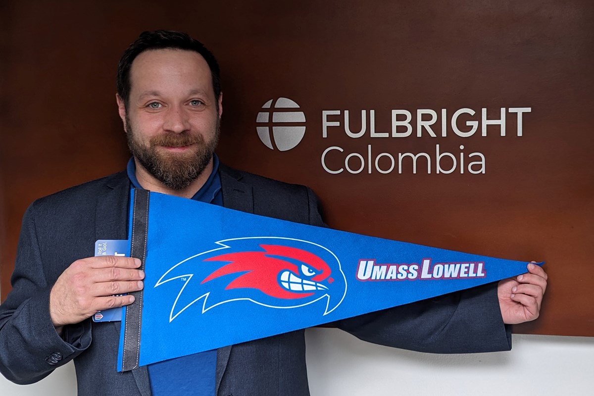 A person with a beard poses for a photo while holding a UMass Lowell pennant.