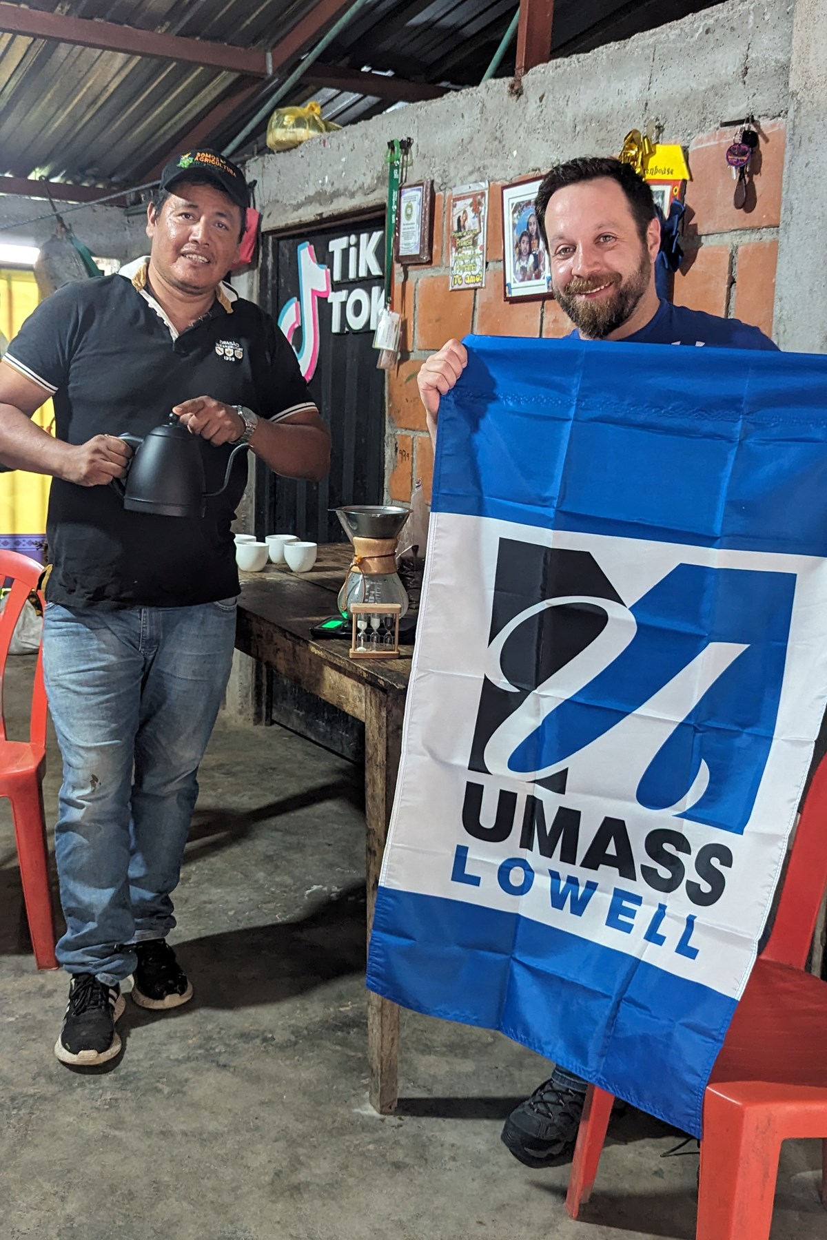A person with a beard holds a UMass Lowell flag while posing for a photo with a person holding a coffee pot.