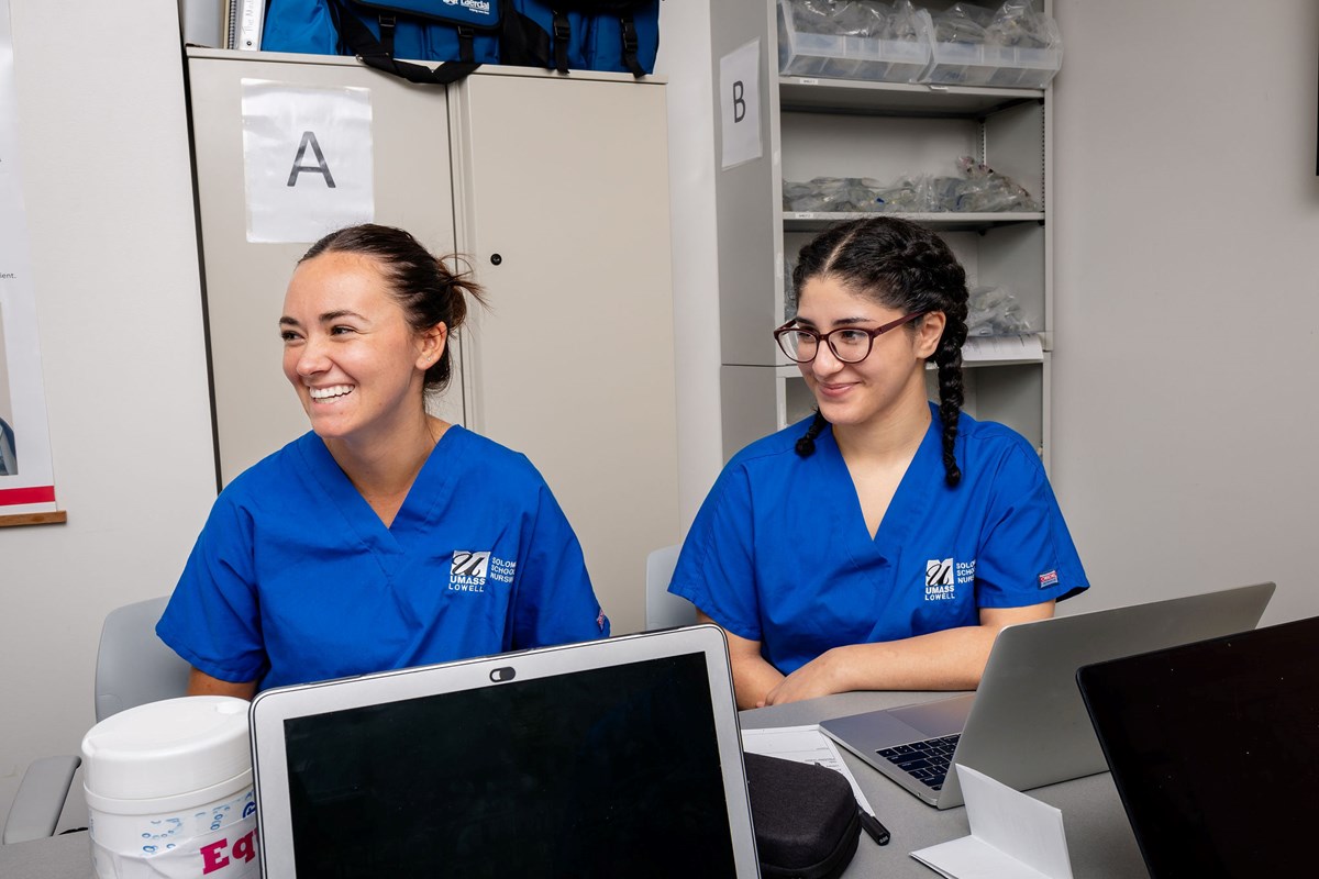 Two nursing students smile as they work at laptops in the UMass Lowell Nursing Simulation Lab.