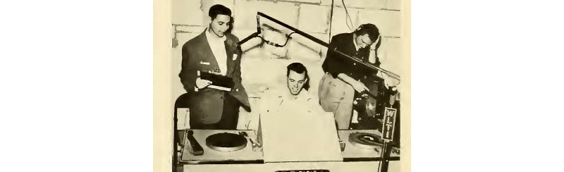 3 students broadcast from a radio station. The desk in front of them has turntables & a microphone.