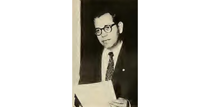 A person in a suit and glasses holds a piece of paper.