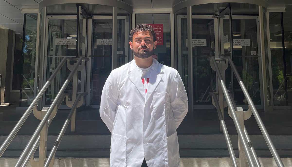 Zachary Kyros stands outside a building wearing a medical coat.