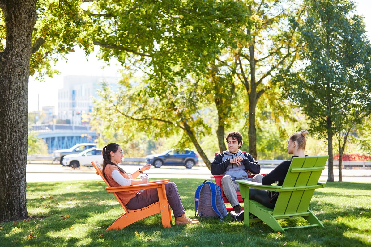 Three students sit in colorful Adirondack chairs under tree on UMass Lowell's North Campus.