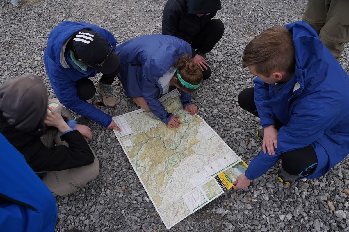 A group of five people look intently at a map on gravel surface.