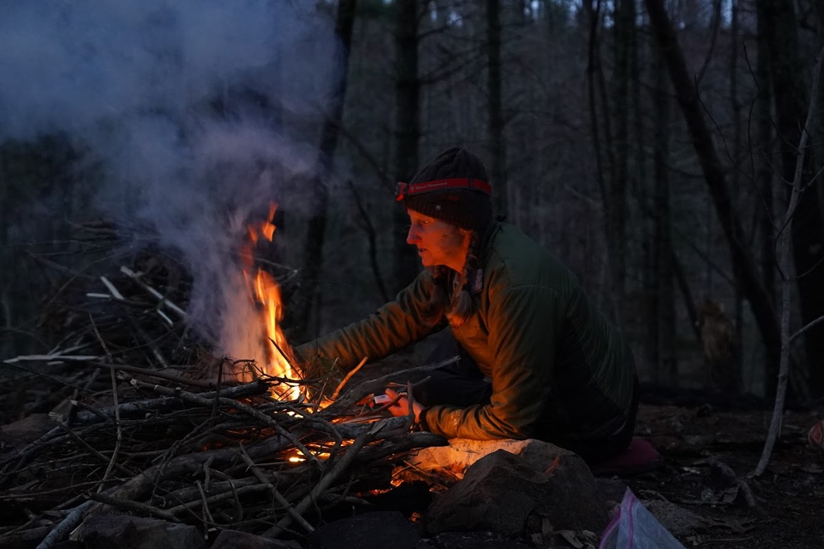 A woman is warmly lit as she reaches across to the side of a small campfire with a pile of unburned sticks.