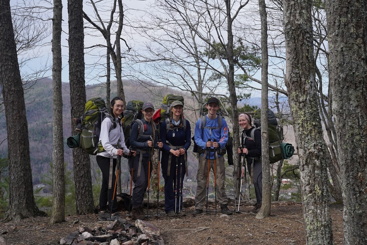 Five hikers stand smiling with backpacks and hiking poles surrounded by bare trees.