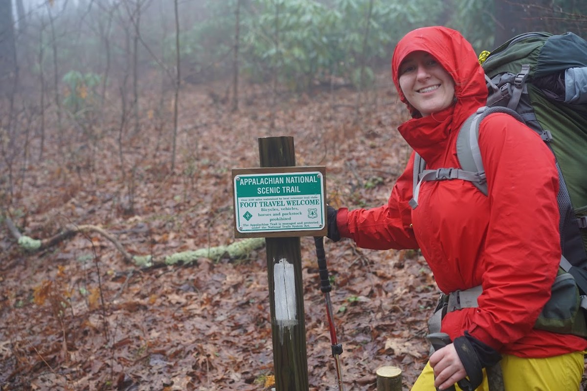 A woman in a bright red coat smiles as she points to a small green Appalachian Trail sign on a post in the woods.