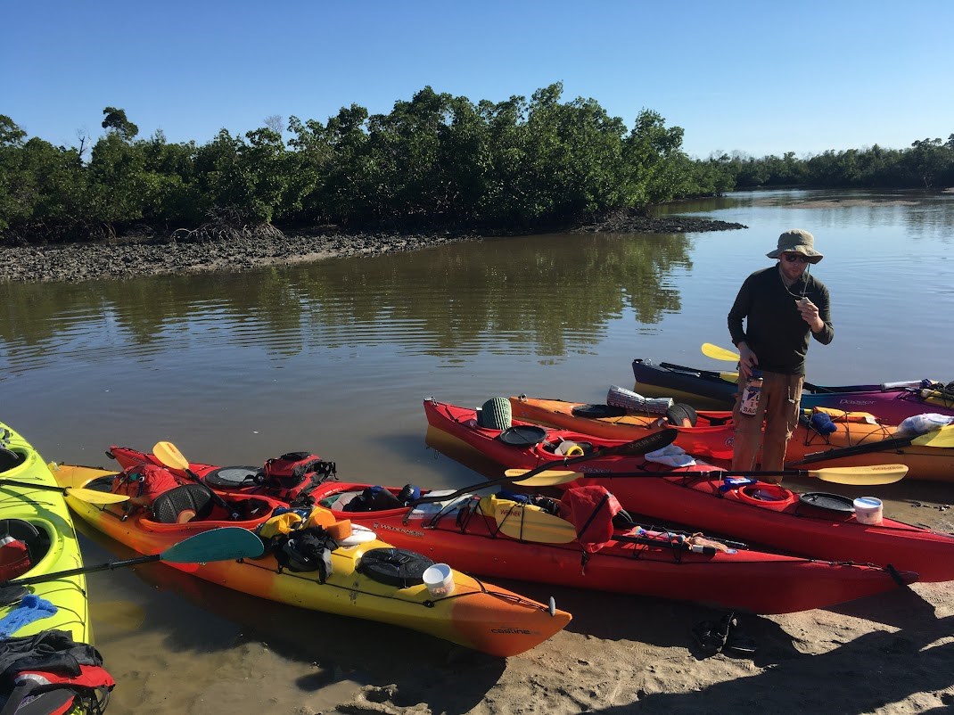 a person stands next to kayaks lined up on the beach with mangroves behind