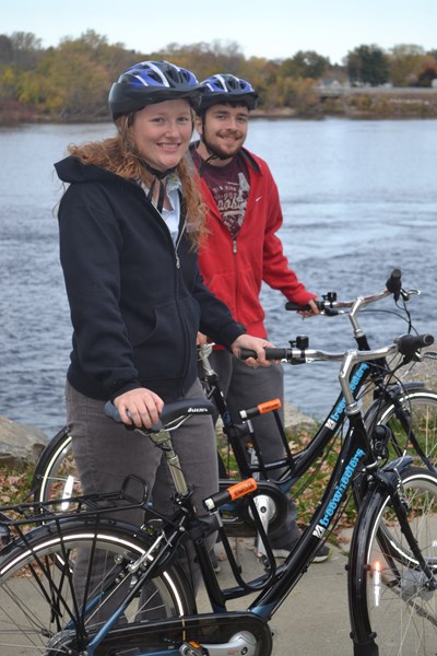 Two students wearing helmets and standing with Free Wheeler bikes in-front of a body of water.